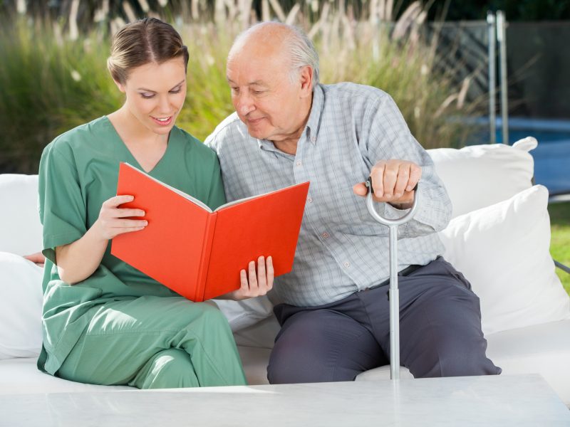 Senior man reading book with female caretaker on couch at nursing home